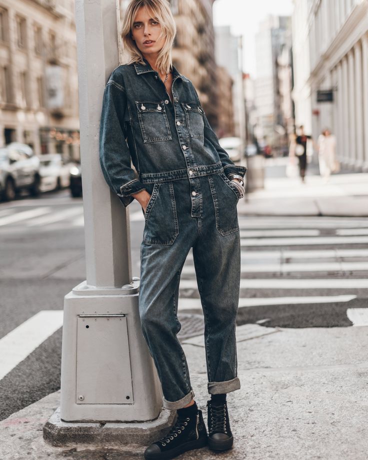a young blonde woman is standing next to a lamppost in a city, wearing a denim jumpsuit and black boots. she is looking at the camera, and the sidewalk is visible in front of her. she has a casual and stylish look.