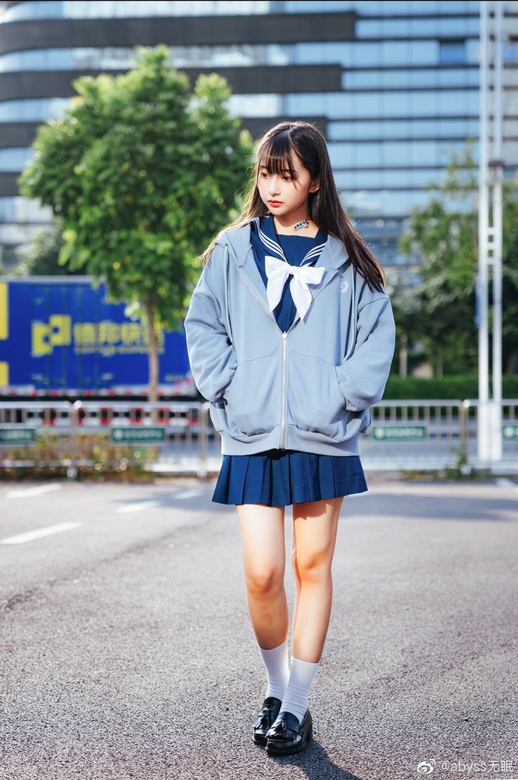 a full body shot of a young asian girl wearing a light blue hoodie over a dark blue sailor uniform with a pleated skirt. she is wearing white socks and black shoes. she is standing on a street in front of a city building. the girl has long dark hair and is looking down with a serious expression. the image is well lit and taken in daylight.