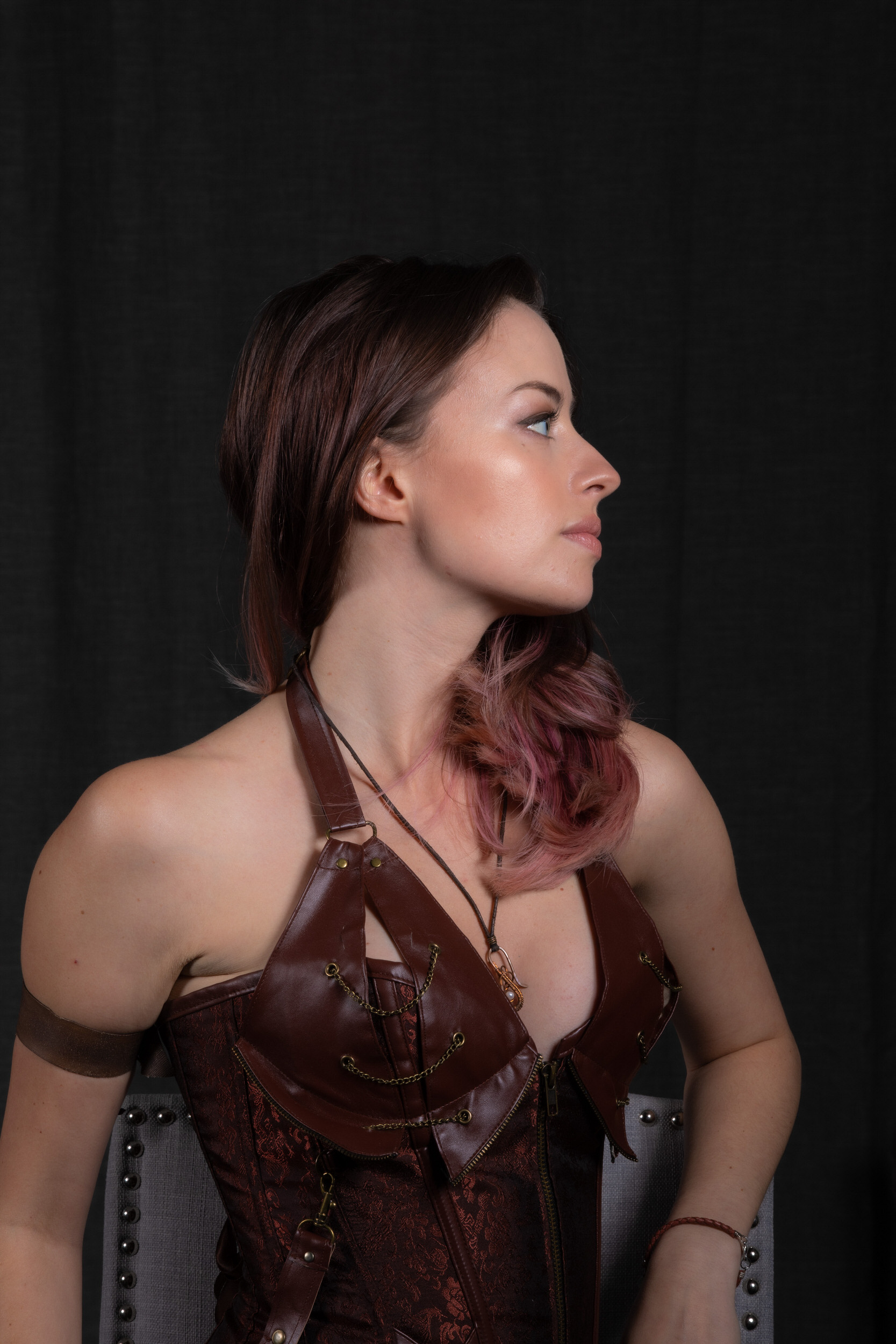 a young woman with pink and brown hair is posing in profile against a black backdrop. she's wearing a brown steampunkinspired corset top with buckles and chains, a brown wrist cuff, and a long necklace with a pendant. the lighting is dramatic, emphasizing her features and the details of her costume.