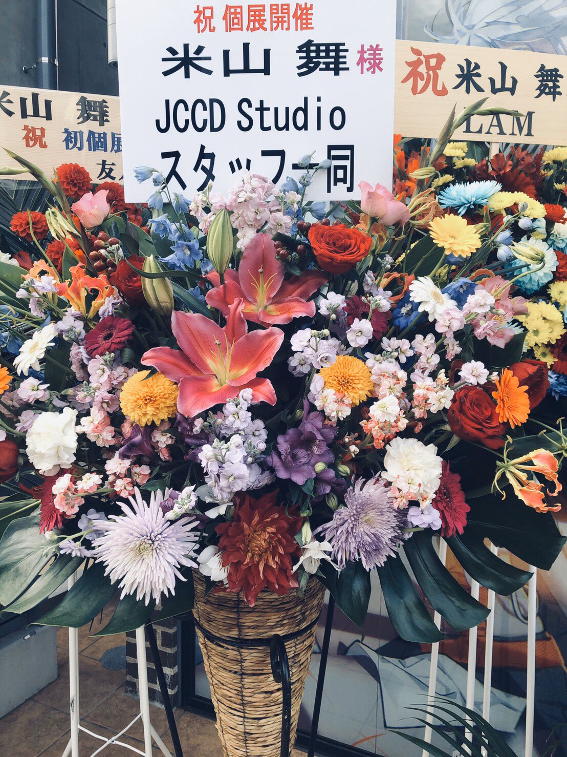 a vibrant floral arrangement celebrating the opening of an art studio, likely owned by an artist named "米山 舞" (yoneyama mai). the arrangement features lilies, roses, and various other flowers in a spectrum of colors, with a congratulatory message written in japanese. the setting appears to be the entrance of the art studio.