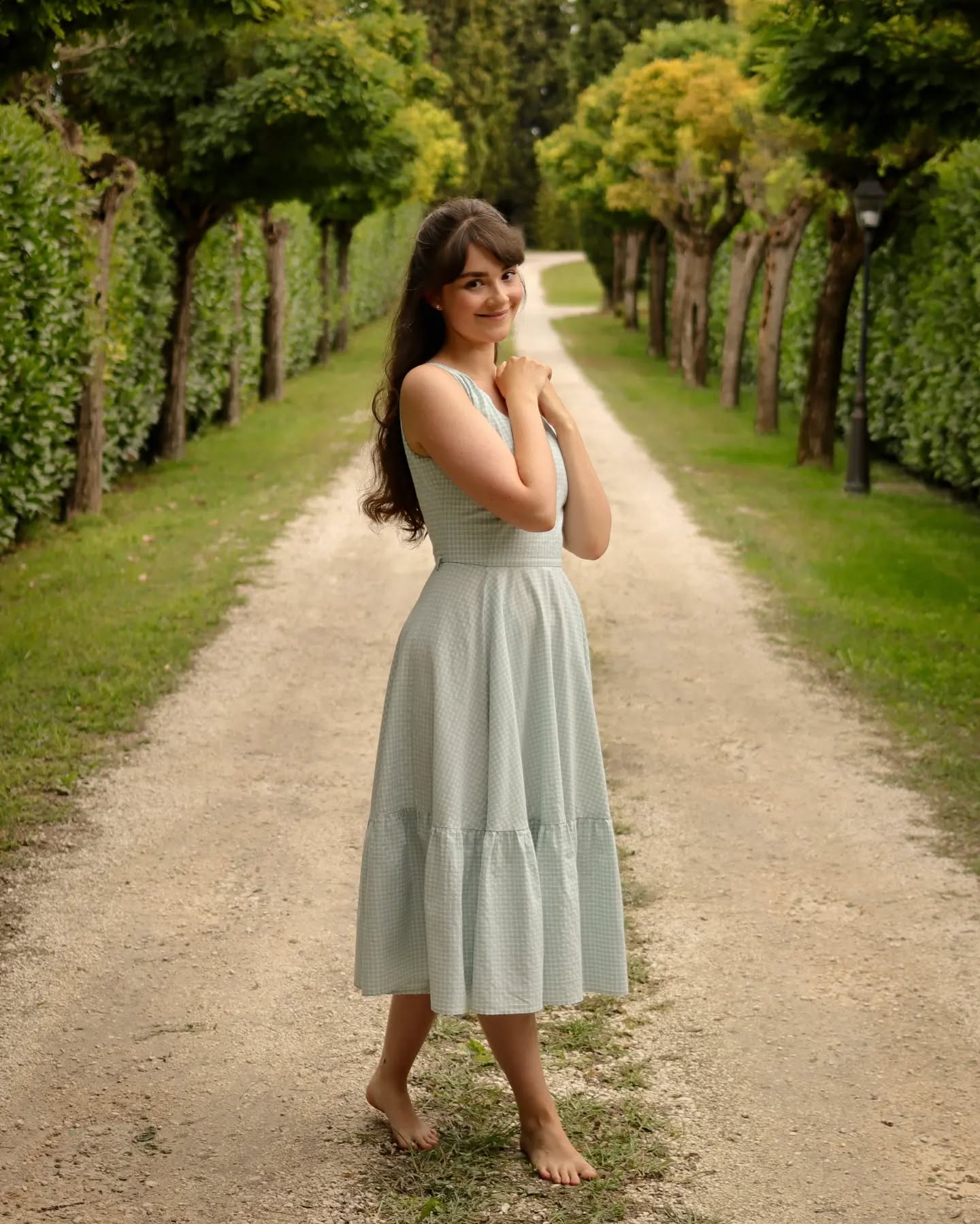 a young woman with long brown hair is standing on a gravel pathway, smiling. she is wearing a light blue gingham midi dress and is barefoot. the pathway is lined with neatly trimmed green hedges and trees, creating a serene and peaceful atmosphere. the overall feeling of the image is one of summer happiness and tranquility.