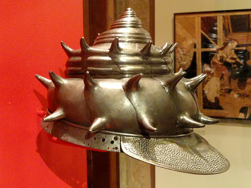 a closeup view of an elaborately crafted silver helmet shaped like a conch shell with sharp spikes protruding from each whorl. the helmet is displayed on a stand against a red wall, with a framed artwork visible in the background.