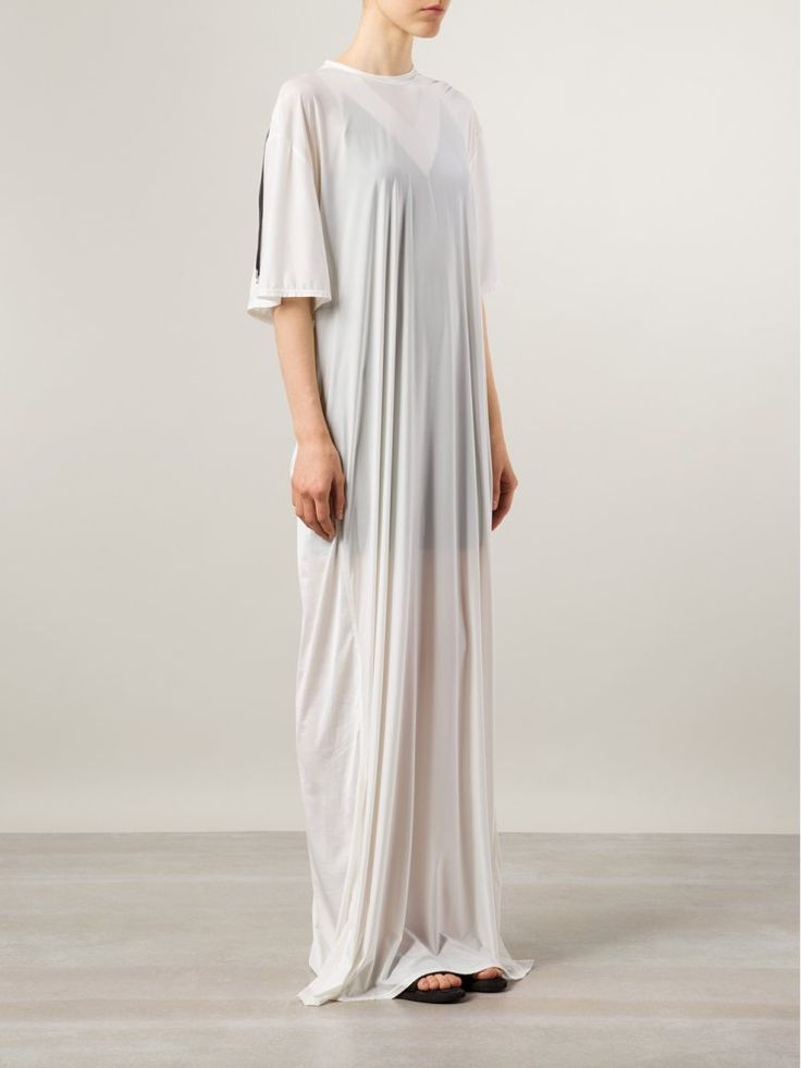 a fulllength studio shot shows a slender, lightskinned model wearing a long, loose, white tshirt style dress that is somewhat sheer and has a black stripe on the left sleeve. the model is facing slightly to the left, but looking straight ahead. the dress drapes to the floor and covers most of her black sandals. the background is a light gray gradient.