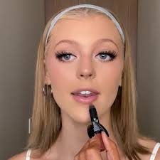 a fairskinned blonde woman with a headband is applying lipstick to her lips. she is holding a tube of lipstick in a black rocketshaped container. she is wearing makeup and looking at the camera. the background is a neutral color.