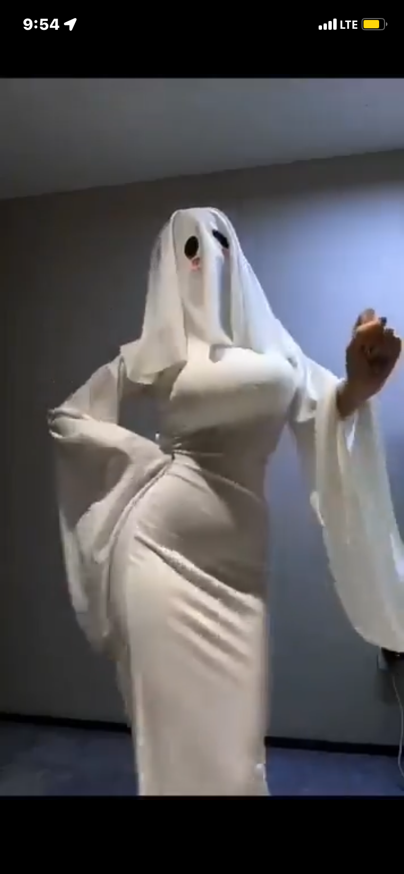 a woman wearing a white ghost costume. she's in a room with white walls, wearing a long white dress and a white sheet over her head. she's holding her hands up in a spooky gesture.