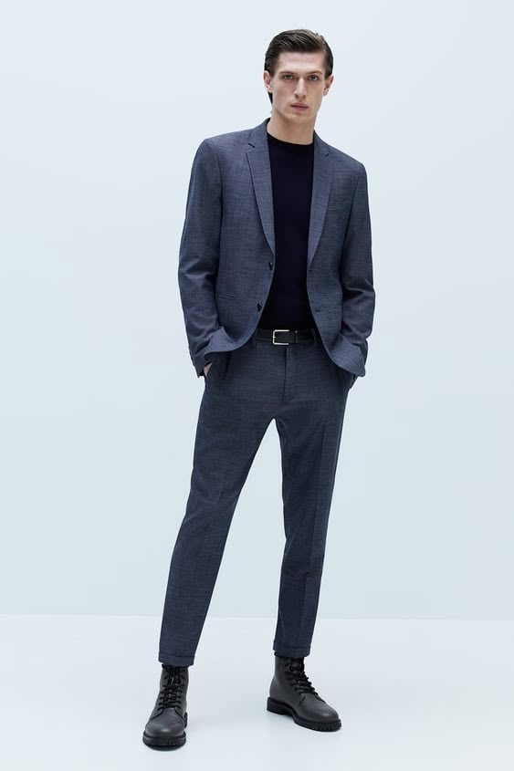a full shot of a man in a suit standing in a studio setting. he is wearing a dark blue blazer and matching pants, a dark blue sweater, a black belt, and black boots. he is facing the camera with his hands in his pockets.