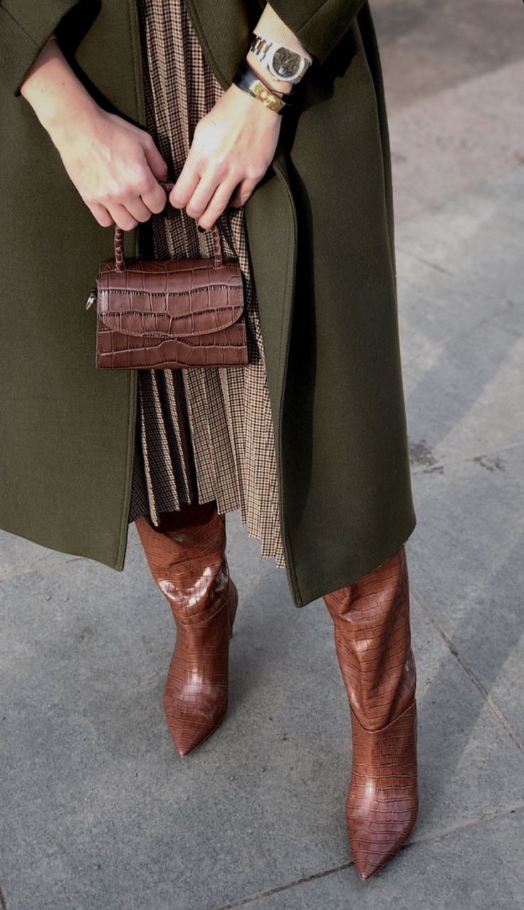 a woman is standing outdoors, showcasing a stylish autumn outfit. she's wearing a long olive green coat over a pleated brown and white skirt. her accessories include brown crocodilepatterned heeled boots, a small brown crocodilepatterned handbag, and a watch on her wrist. the overall style is chic and sophisticated, fitting for fall weather. the image is welllit and shows the details of her outfit and accessories clearly.