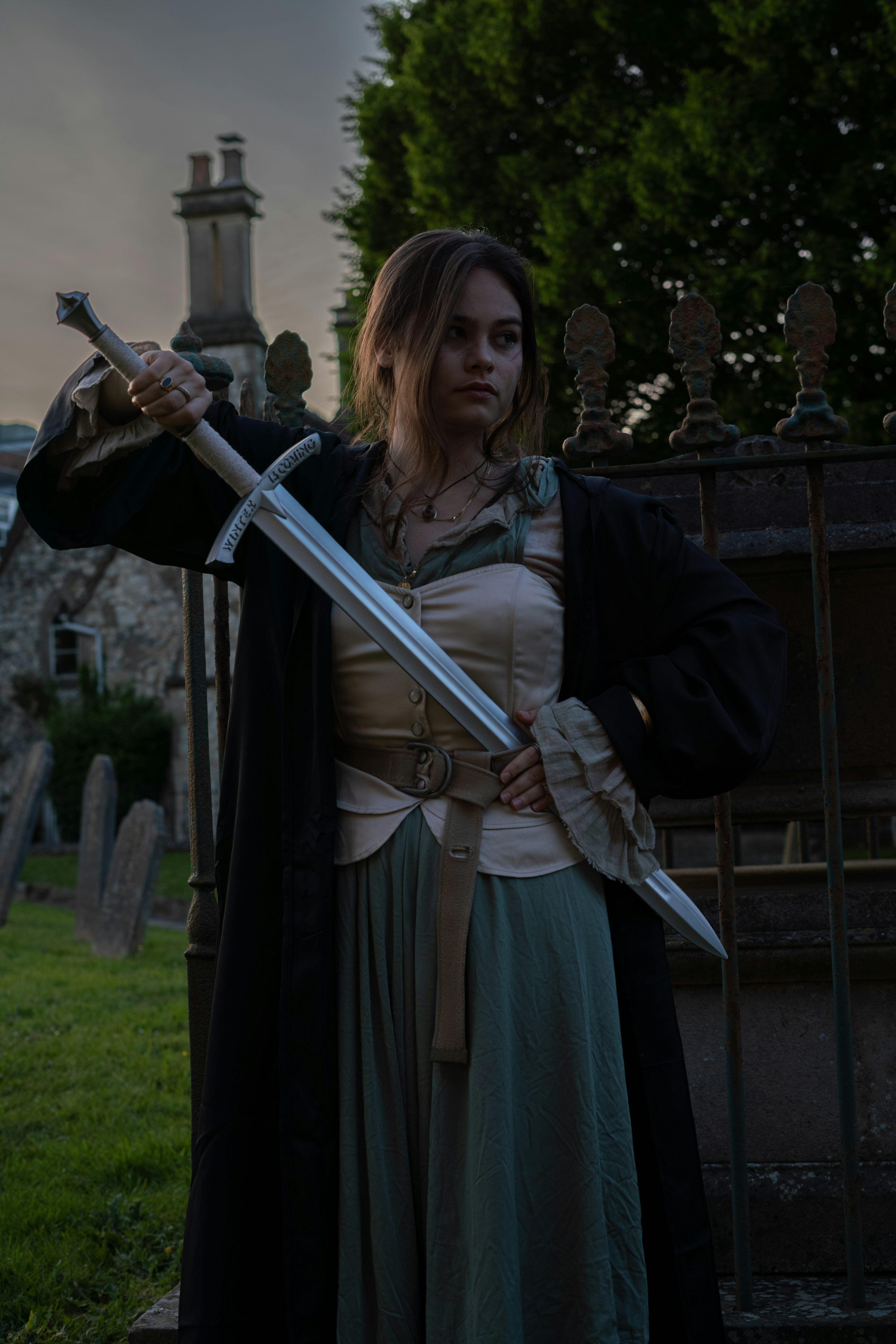 a young woman, dressed in a medievalstyle costume, stands in a graveyard at dusk. she is holding a sword, her expression is serious and determined. the scene is dark and atmospheric, with a gothic feel. the background features a stone fence, trees and tombstones. she is wearing a long tealcolored dress under a dark cloak or robe, and a beige bodice. the sword she holds appears to be a replica of a medieval or fantasy weapon.  the overall mood is one of mystery and power.