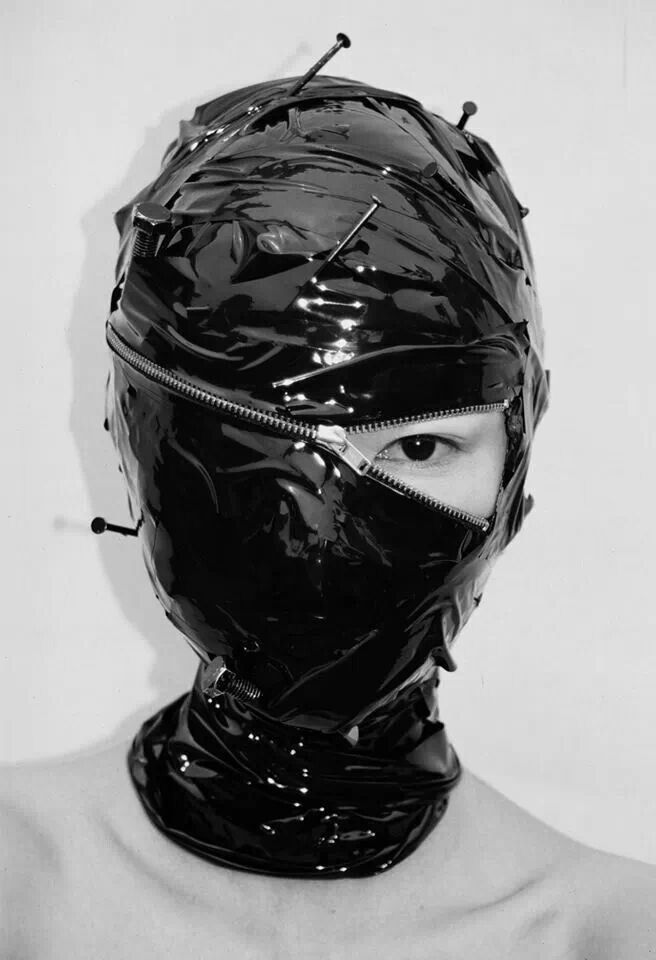 a woman wearing a black mask made from plastic,  with a zipper down the middle and a single eye hole on the left side. the mask is covered in pins. the woman's skin is visible around the mask's edges. the background is plain and white.