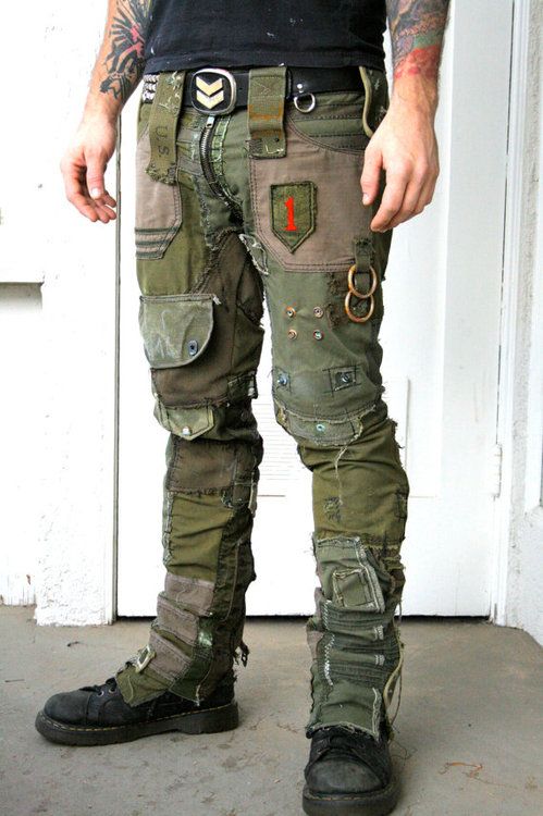 a man wearing a black shirt, brown belt with a silver buckle, and patched up green camouflage pants with the same pattern on his boots, poses for a photo in a room.  the man is wearing a distressed look.