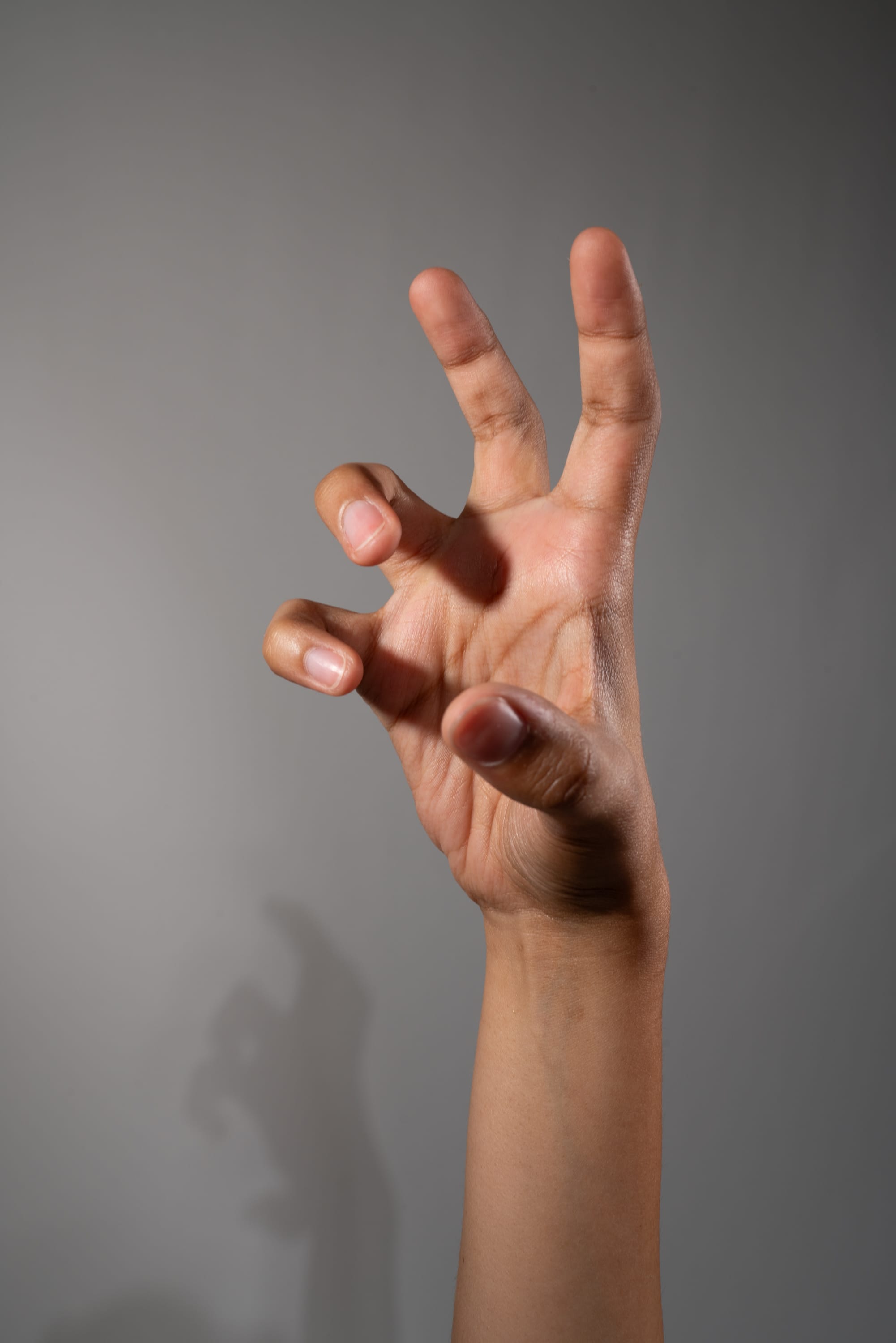 a single hand is extended against a neutral gray background, fingers curled in a clawlike gesture. the hand is lit from the side, creating a distinct shadow on the wall behind.