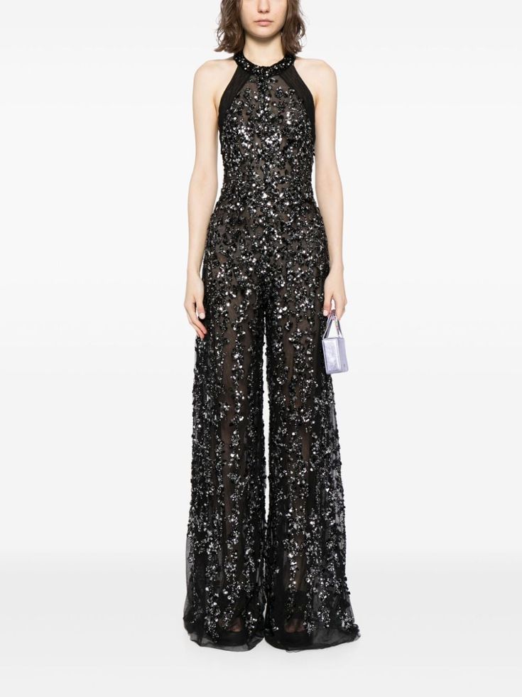a woman wearing a black sequined halter top jumpsuit with wide legs. the jumpsuit is floor length and features a sparkling silver detail throughout. she is holding a small silver clutch bag and accessorizing with a silver necklace. she is posing in a studio setting against a white background.