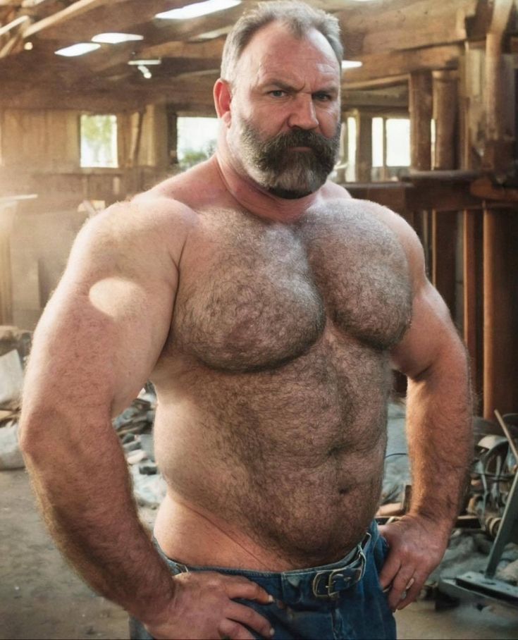 a hairy, muscular man with a thick beard stands in a workshop, looking directly at the camera. he has his hands on his hips, and his chest and arms are on full display. the man appears to be very strong and fit, with a muscular build and a defined six pack. he is wearing a pair of blue jeans and has a lot of body hair, especially on his chest and arms. the man's expression is serious and intense.