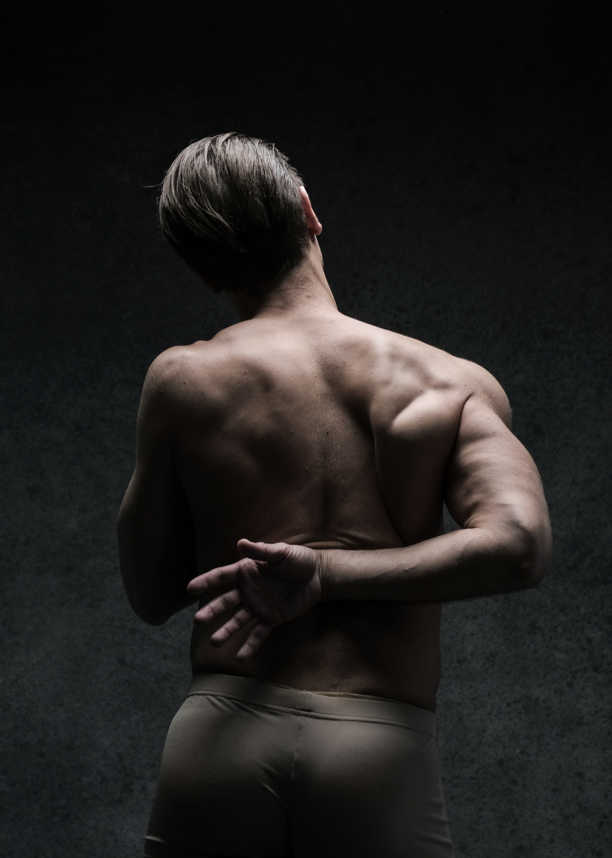 the image showcases a muscular man from the back, his upper body bare and illuminated by studio lighting against a dark backdrop. his pose, with his head tilted back and one arm reaching behind him, emphasizes the definition and strength of his back muscles. the monochrome palette further accentuates the play of light and shadow on his physique.