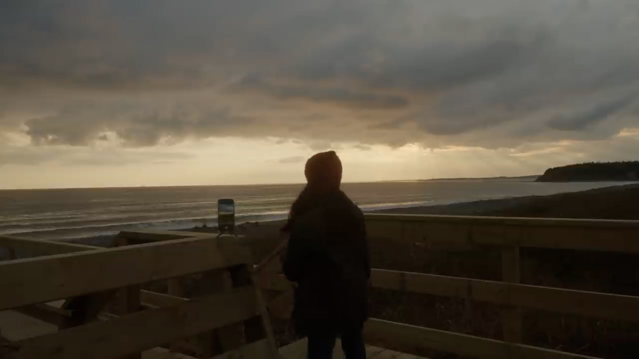 a woman stands on a wooden boardwalk, her back to the camera, capturing a picture of the ocean with her phone on a tripod. the sun is setting over the water, casting a warm glow on the clouds. the sea is slightly rough, and a rocky headland is visible in the distance.  the overall lighting is muted and overcast.