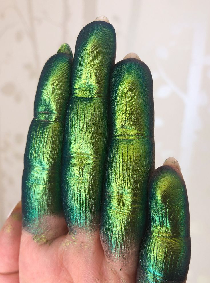 a hand with four fingers painted with a green iridescent eyeshadow. the eyeshadow has a shimmery, metallic finish. the hand is held up against a white background.