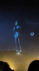 a silhouette of a person performing on stage at a concert. the stage is lit with bright lights, and the crowd is visible in the distance. the image captures the energy and excitement of a live music event.