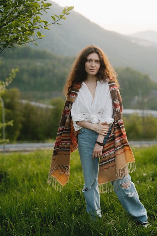 a young woman with long brown curly hair stands in a grassy field with mountains in the background. she is wearing a white shirt, distressed light blue jeans, and a large, colorful patterned poncho or shawl. the setting is outdoors in nature, with the light suggesting it's daytime. her expression is calm and serene.  the overall aesthetic is bohemian or hippieinspired.