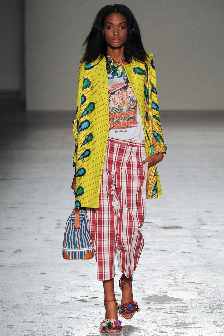 a runway model walks down the catwalk wearing a vibrant yellow coat with a peacocklike print, a white graphic tee, and red and white checkered cropped pants. the model accessorizes with a striped tote bag and flowerembellished heels. her dark hair is styled in loose waves. the overall look is bold, colorful, and eclectic, showcasing a mix of prints and textures. the background is a light gray runway.