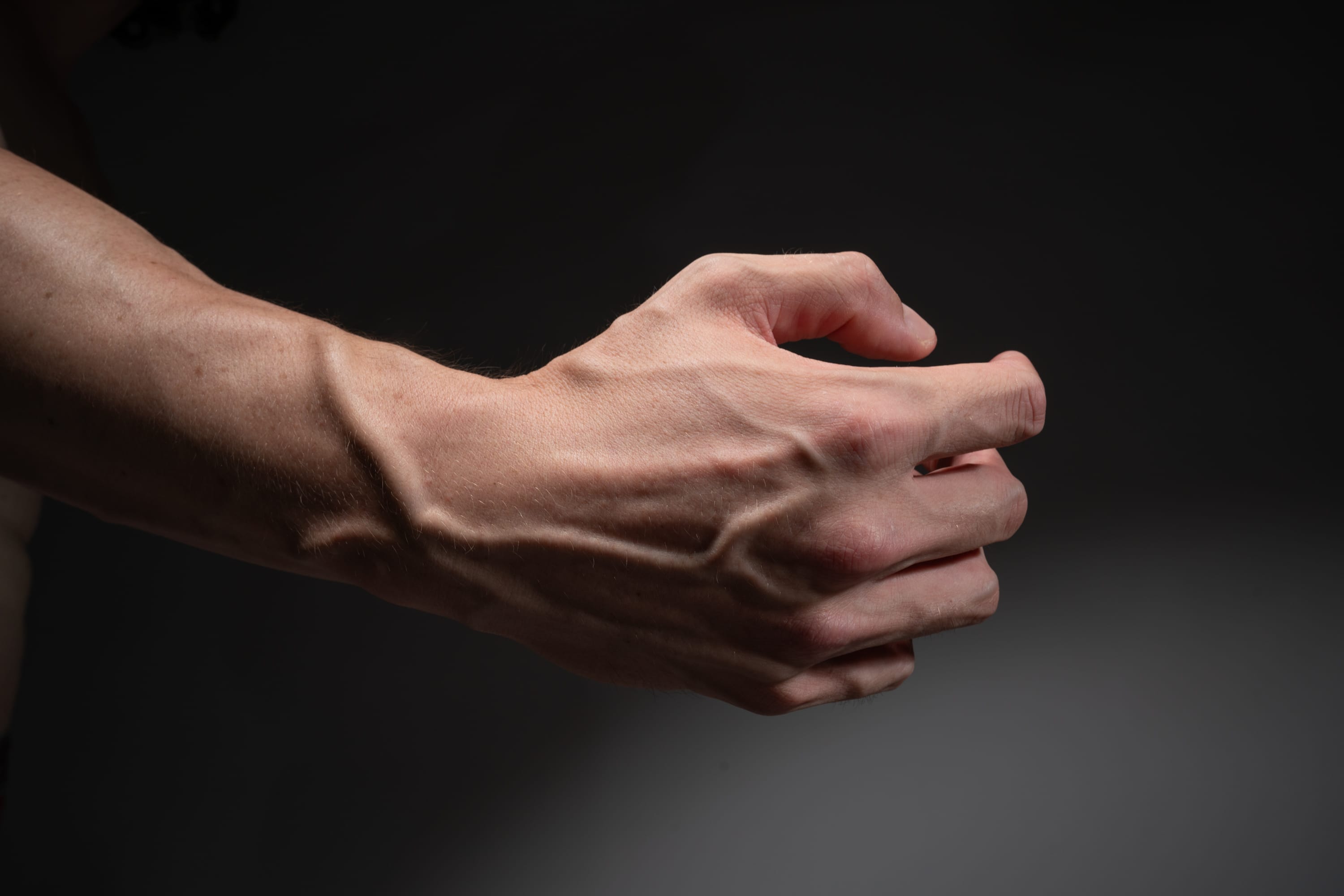 a closeup side view of a muscular man's arm and hand, showcasing prominent veins and tendons. the lighting is soft and diffused, highlighting the texture and contours of the skin. the background is a dark gray, creating a strong contrast and emphasizing the subject.