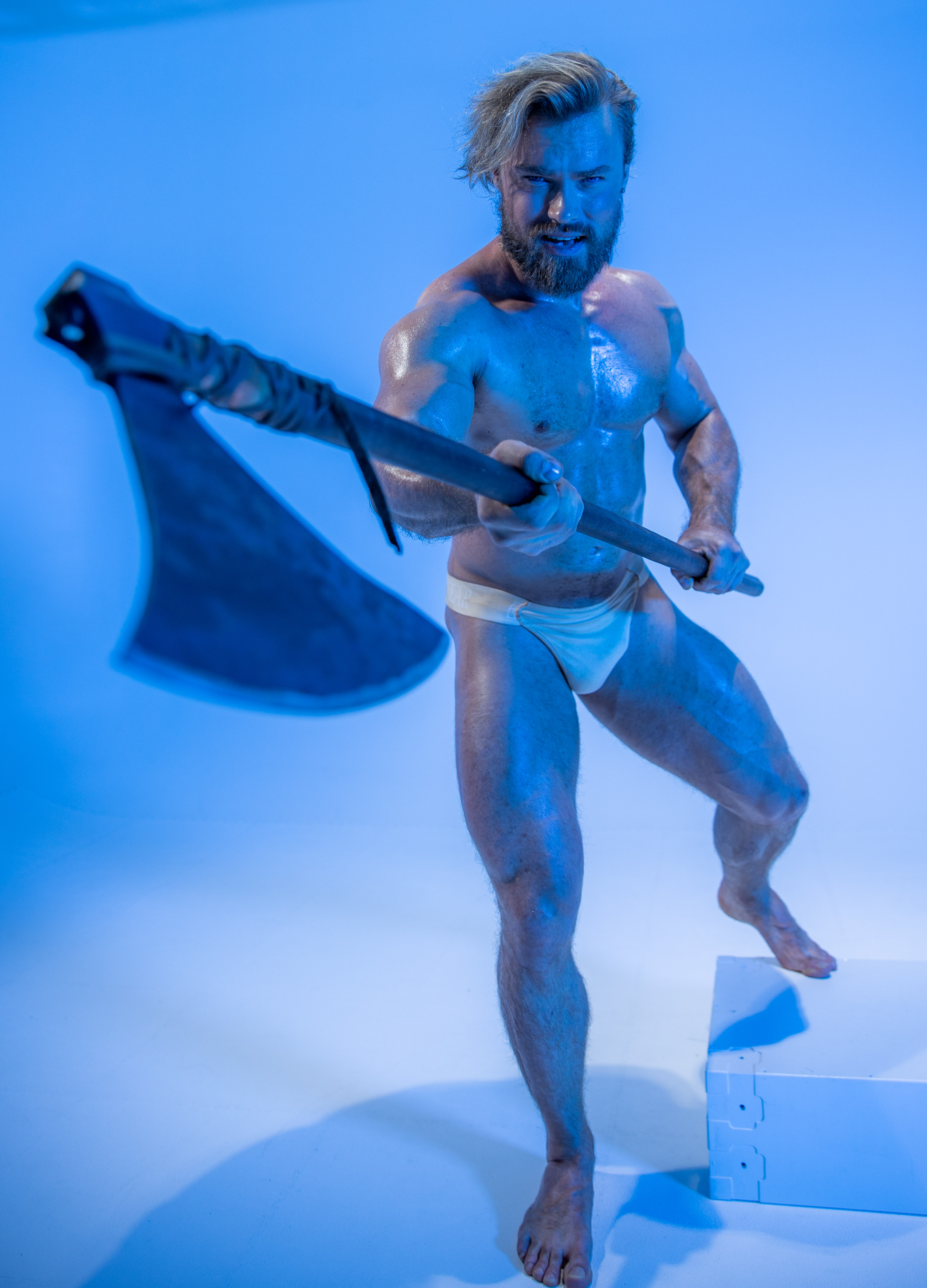 a muscular, shirtless man with long blonde hair and beard, styled as a viking warrior, poses against a vibrant blue background. he grips a large, doubleedged axe with a determined expression, its blade prominent in the foreground. the low camera angle emphasizes his physique and the weapon's imposing size.  he stands on a white platform. the lighting accentuates his muscles, creating a dramatic and powerful image.