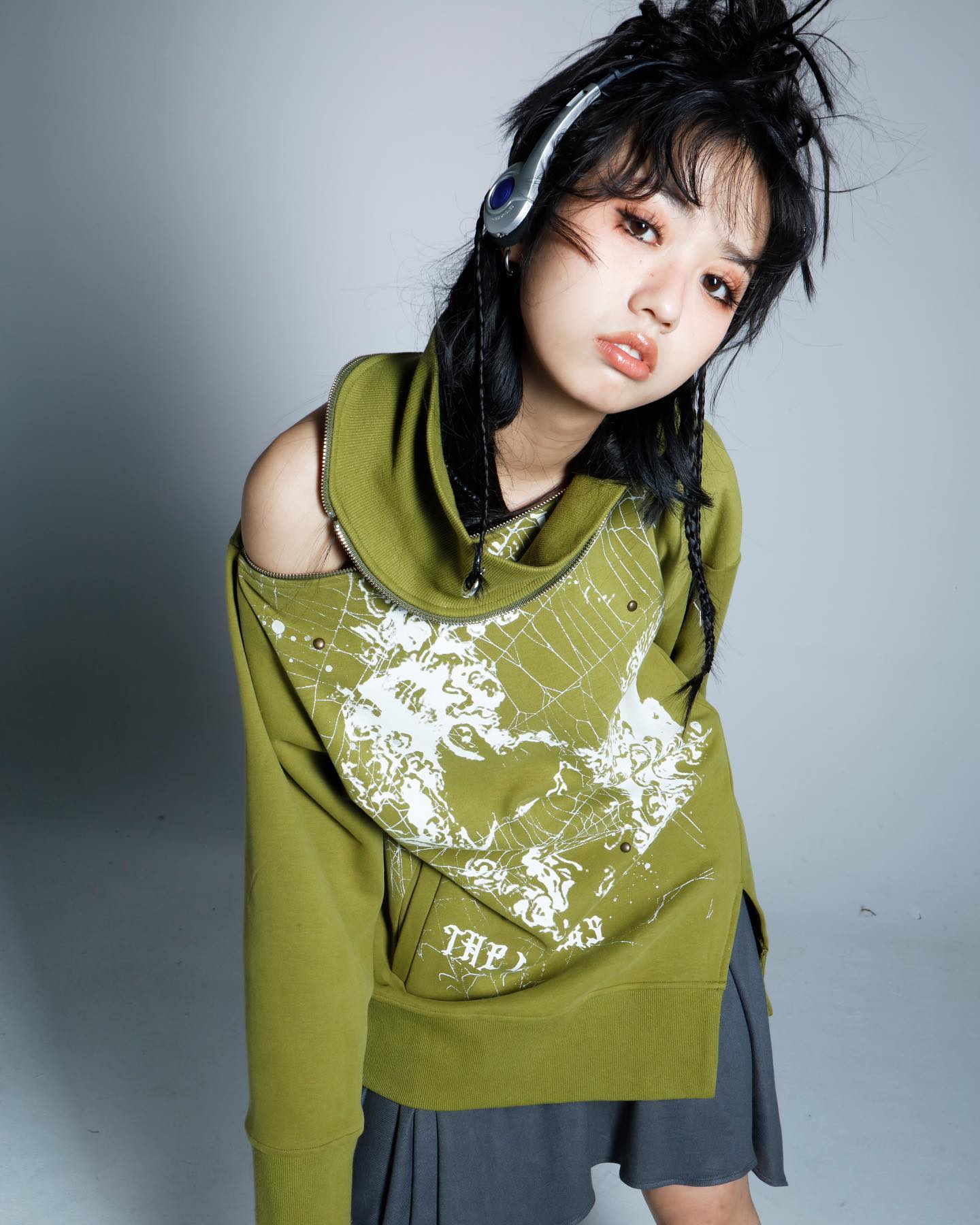a young woman with dark hair styled in braids and wearing headphones is posing in a stylish olive green hoodie with a graphic skull and spiderweb print. the hoodie has an offtheshoulder design and a high neck. she is also wearing a grey pleated skirt. the background is a simple grey. the overall style is edgy, stylish, and streetwearinspired.