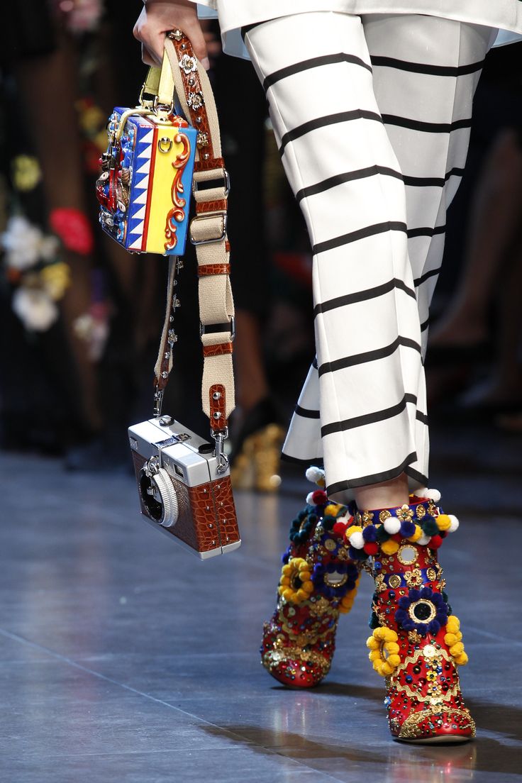 closeup shot of a model's legs at a fashion show, showcasing dolce & gabbana's collection. the model is wearing black and white striped pants, colorful pompom embellished boots, and carrying a unique handbag designed as a camera and a box bag. the image emphasizes the intricate details of the accessories and the model's stylish outfit. the background is blurred, focusing attention on the model's attire and accessories.