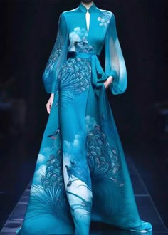 a model in a long, flowing blue dress with peacock print stands on a runway. the dress has long, flowing sleeves and a fitted bodice. she is posed in a striking pose, showcasing the elegance of the gown. the dress is a beautiful and stylish piece of clothing.