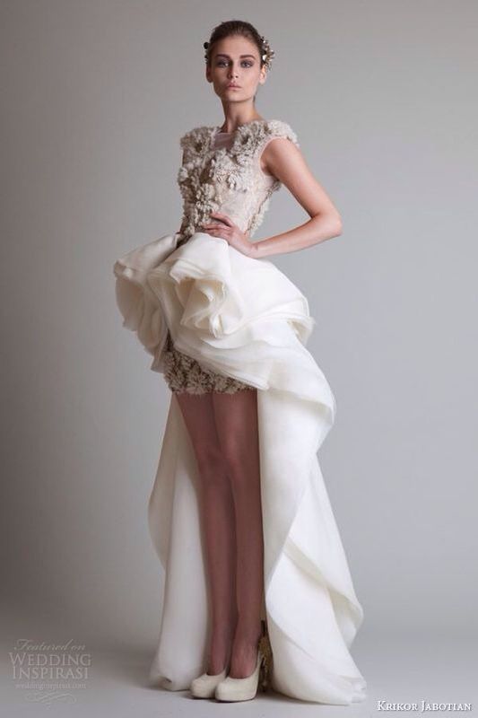 a beautiful bride model wearing a highlow wedding dress with ruffles and flower embellishments. the dress has a short front and a long train. the model is posing in a studio with a white background. she is wearing her hair up and has a natural makeup look.
