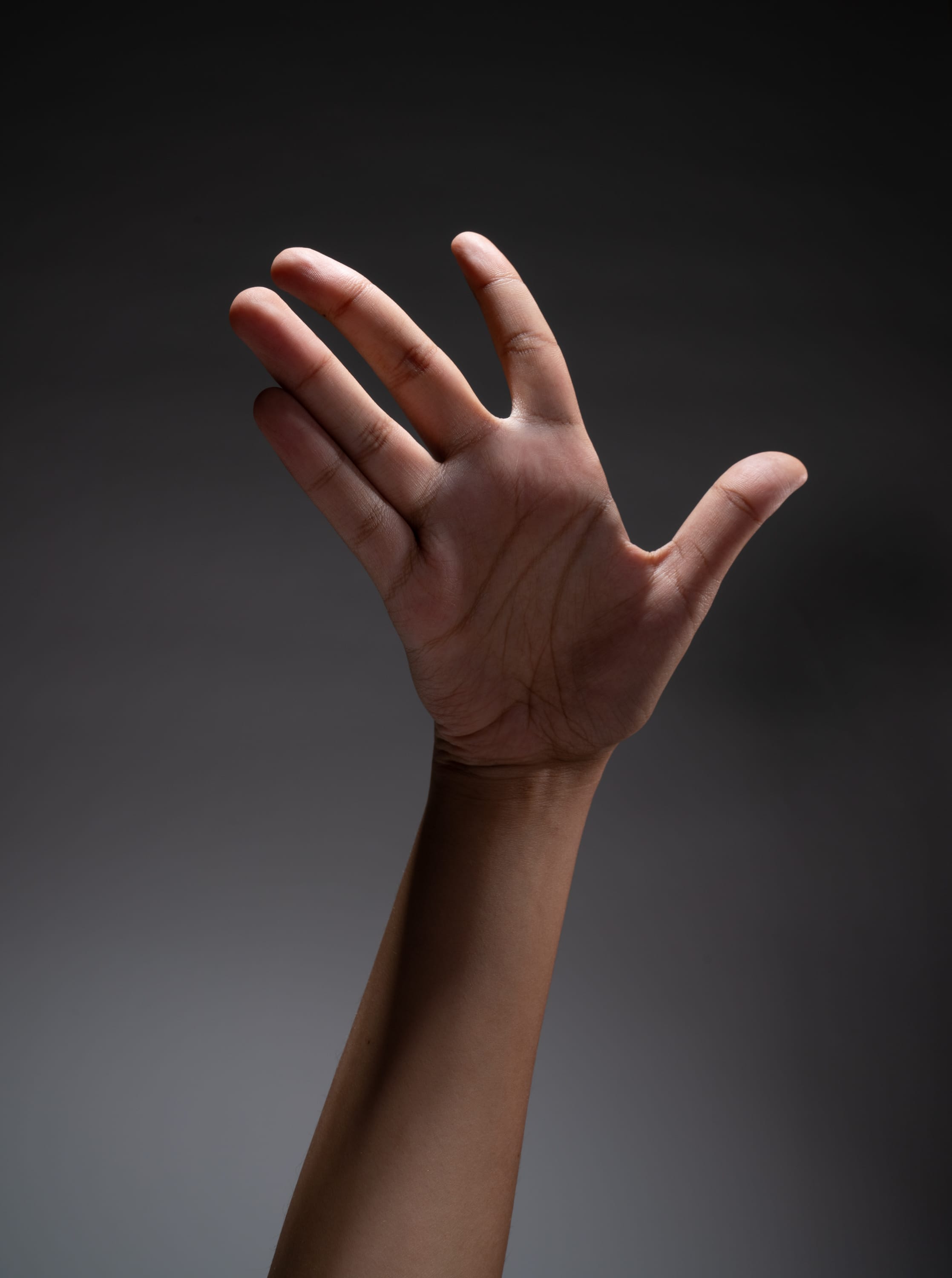 a hand is extended upward against a dark gray background, illuminated by a spotlight. the hand is reaching out with all fingers extended except the thumb, creating a dynamic and expressive gesture.
