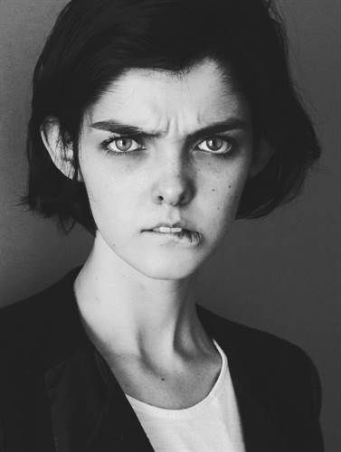 a black and white portrait of a young woman with a serious expression on her face, looking directly at the camera. she has short, dark hair and is wearing a black jacket and a white tshirt. the image has a closeup perspective and captures her sharp features and intense gaze. the lighting is soft and creates a moody atmosphere.