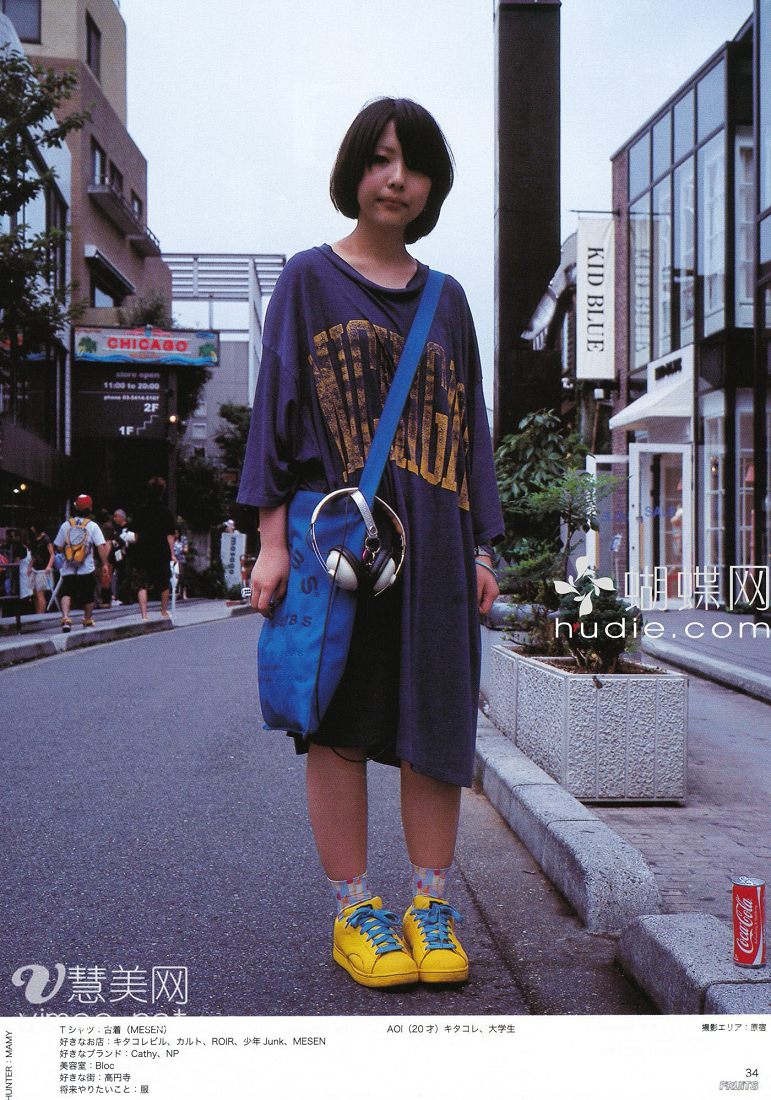 a young woman in japan poses for a street style photo for a magazine.  she is wearing an oversized blue tshirt, black biker shorts, and yellow sneakers.  she is carrying a blue tote bag with headphones attached and stands on a sidewalk next to a coca cola vending machine. the text on the magazine page lists her age, occupation, favorite shops, brands, and hobbies.