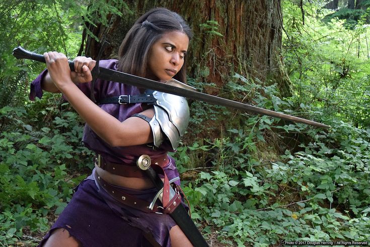 a woman warrior dressed in leather and armor, with a sword in hand, stands in a forest. she looks determined, ready to defend herself. the image has a fantasy setting. the background is a lush green forest.