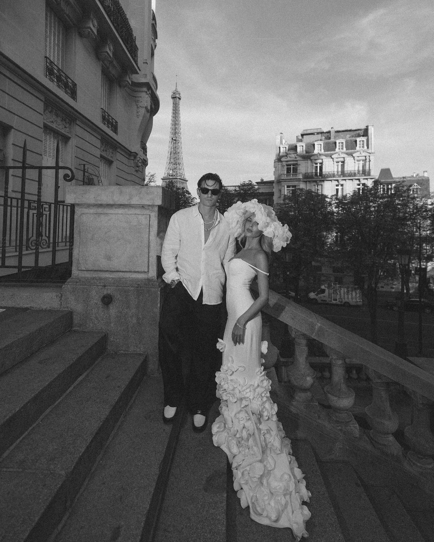 a couple poses for a photo in front of the eiffel tower in paris. the man is wearing a suit and the woman is wearing a white dress with a large hat. they are standing on a set of stone steps. the photo is in black and white.