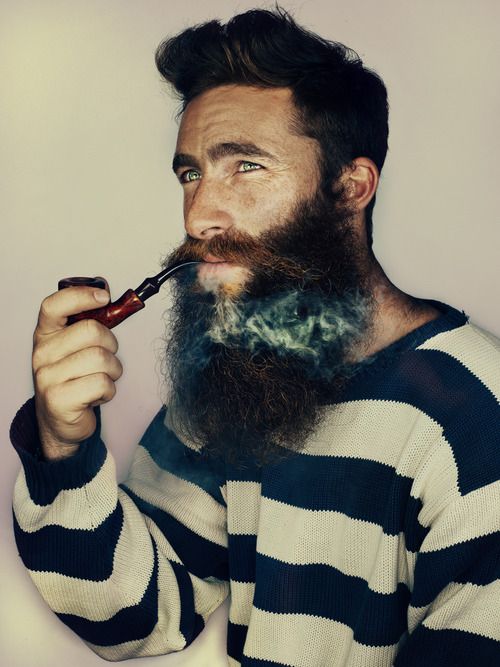 a man with a long, thick beard is smoking a pipe. he is wearing a striped sweater and is looking away from the camera. there is smoke coming out of his pipe.