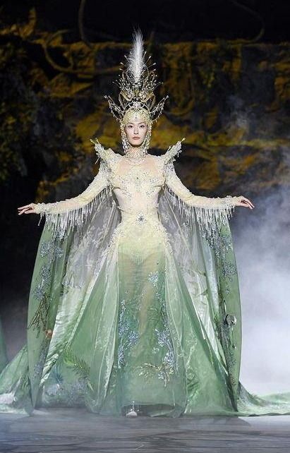 a model walks the runway in a stunning green gown with a delicate and ethereal design. the dress is a sheer fabric, embellished with delicate details and intricate beading. the model wears a crown and jewels that add to the mystical and magical air of the look. the light catches the fabric beautifully, making the gown look like it's flowing and shimmering. the model's pose and expression are full of confidence and grace. the overall look is one of beauty, elegance, and enchantment.