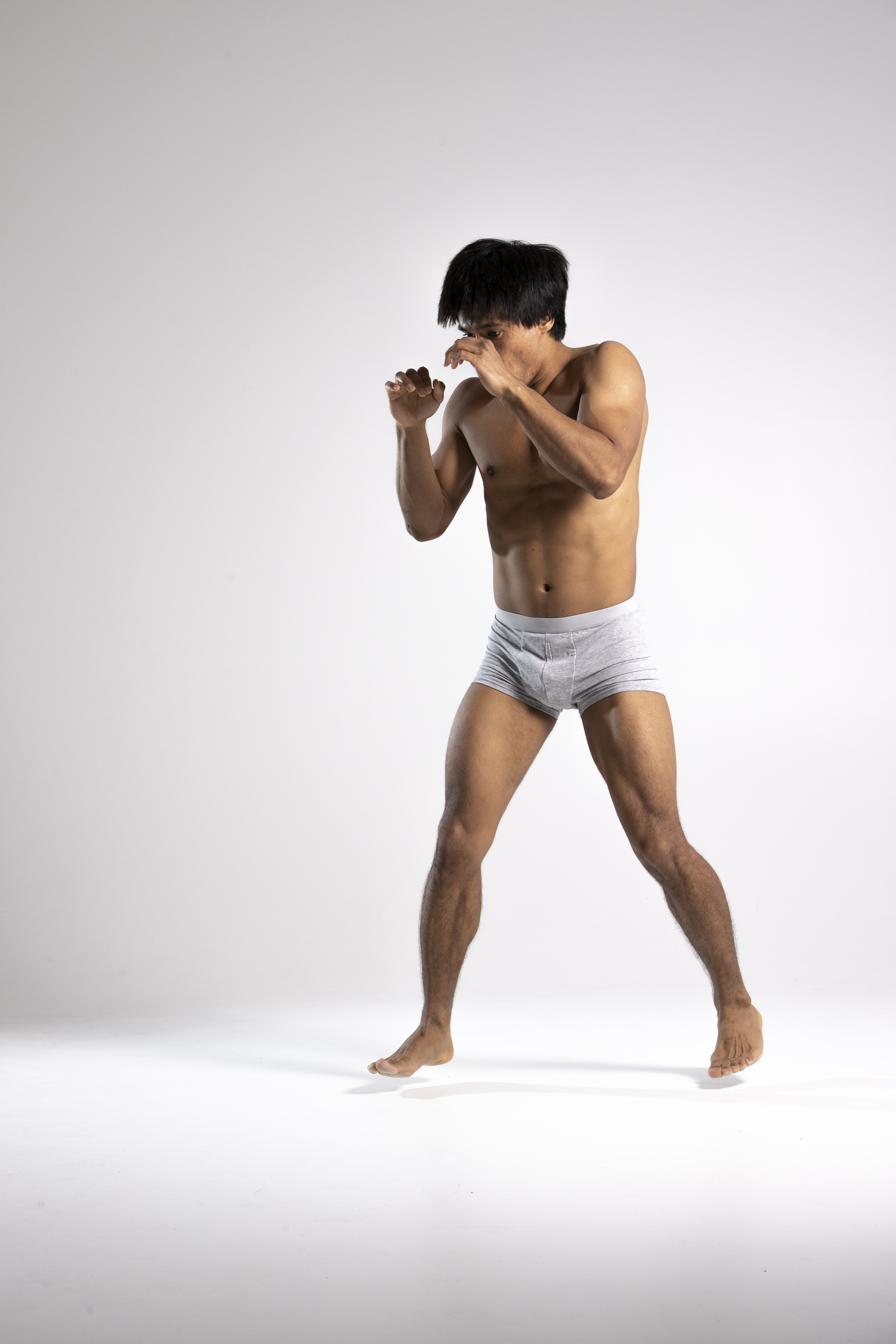 a muscular, barechested asian man in gray boxer shorts is posing in a defensive boxing stance against a gray background in a studio shot. his fists are raised in front of his face, and he is looking forward with a focused expression. his body is toned and athletic.