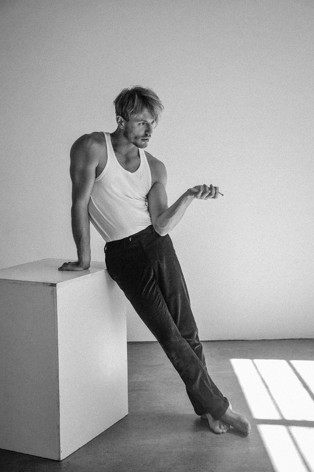 a man in a tank top and black pants is sitting on a white cube in a studio setting. he has his legs crossed and is looking off to the side. the photo is in black and white.