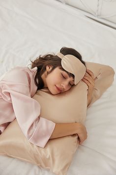 a woman with long brown hair is sleeping peacefully on her side on a bed with white sheets. she is wearing a pink pajama top and an eye mask, and her arms are wrapped around a long, rectangular pillow. the overall mood of the image is calm, serene, and inviting.