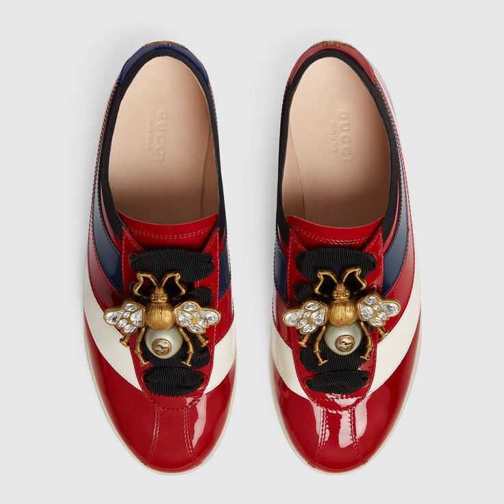 a pair of women's gucci sneakers are displayed against a light gray background. the shoes are red patent leather with blue and white stripes, and feature a bee embellishment with crystals and pearls on the laces. the gucci logo is visible on the insole. the shoes are shown from a topdown perspective, with one shoe slightly angled to the left and the other to the right.