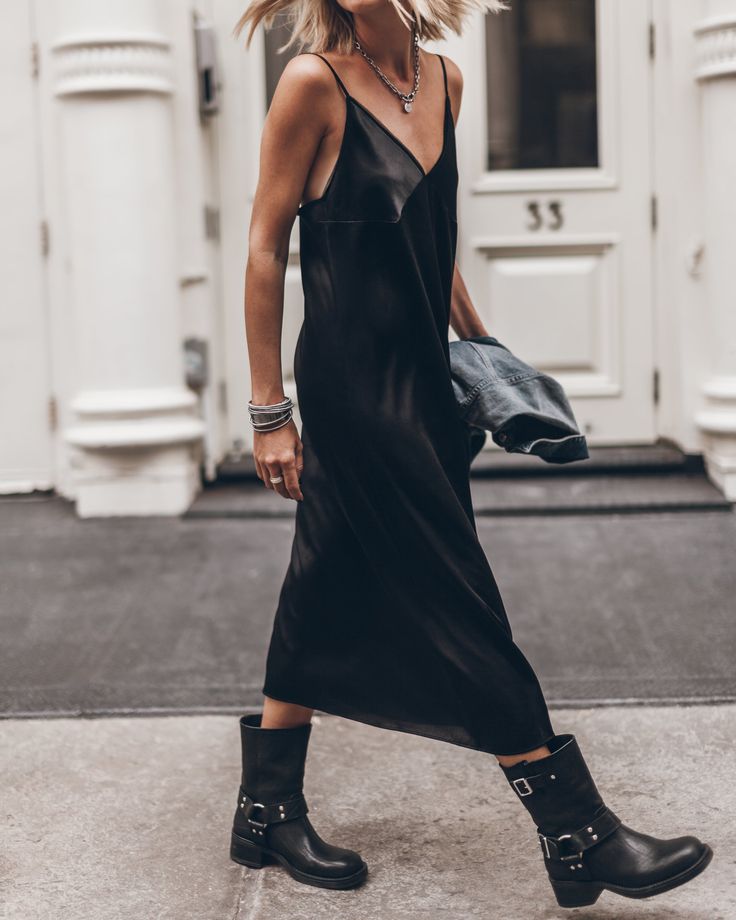 a woman in a black slip dress and black boots walks on a city sidewalk. she is looking to the right of the frame.  the dress is long and flowy, and the boots are chunky and have buckles.  the image has a stylish and casual vibe.