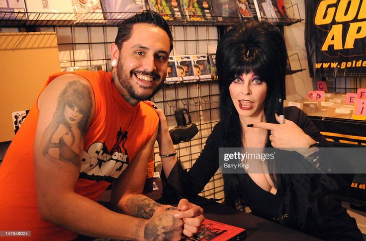 a man with a black tattoo of a woman on his left arm wearing an orange tshirt, poses with a woman with dark hair and dark clothing. she has a large smile and is pointing to her right hand, where the man has his left hand. the man also has a big smile on his face. they are both sitting together in a professional photo setting with a black background, and an assortment of items in the background on shelves that are partially obscured by the people in the image.