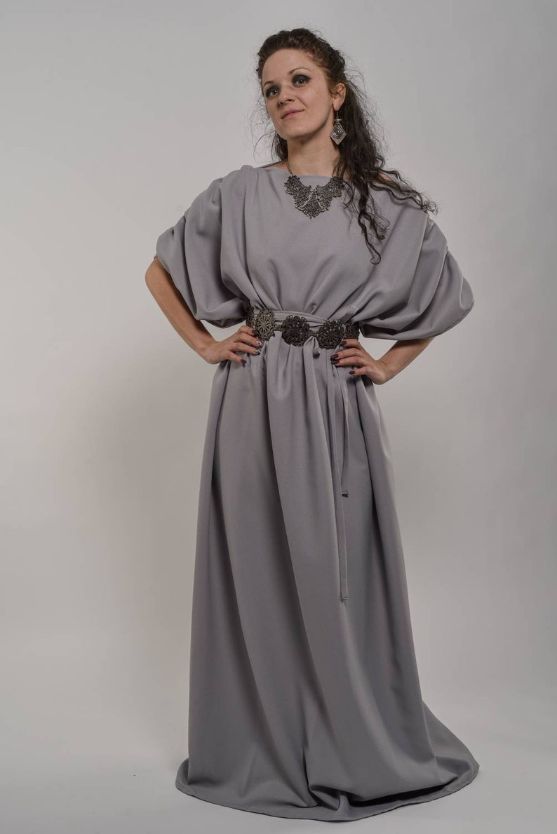 a fulllength studio shot of a woman wearing a long, flowing grey dress. the dress has loose, puffed sleeves and a simple, fitted waist. she is wearing a dark necklace, earrings and a dark patterned belt. the dress falls gracefully to the floor.  the woman has dark curly hair and a contemplative expression. the background is a plain light grey.