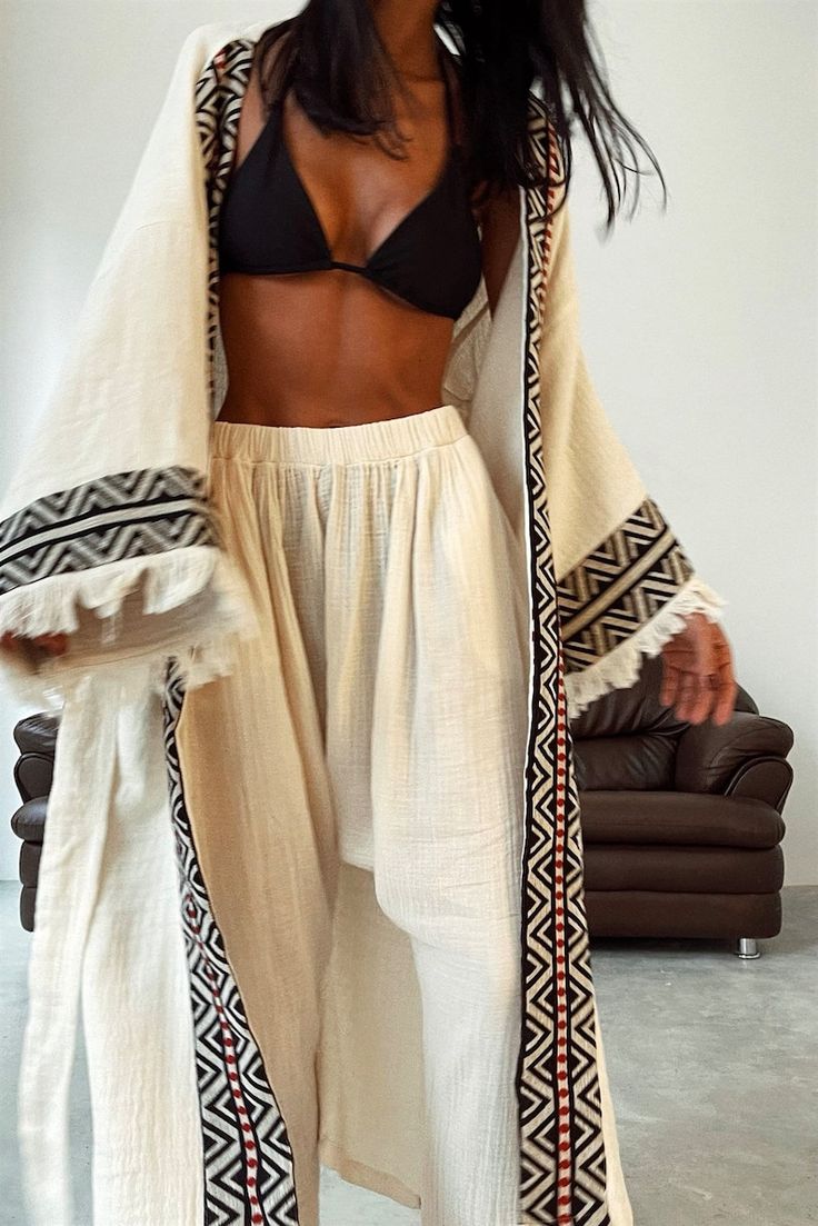 a woman is showcasing a stylish offwhite kimono robe and matching palazzo pants. the robe features black and red geometric patterns along its edges.  she is also wearing a black triangle bikini top underneath. the model is posed casually, indoors, against a simple background featuring a brown leather sofa. the overall style is relaxed, bohemian, and beachy.