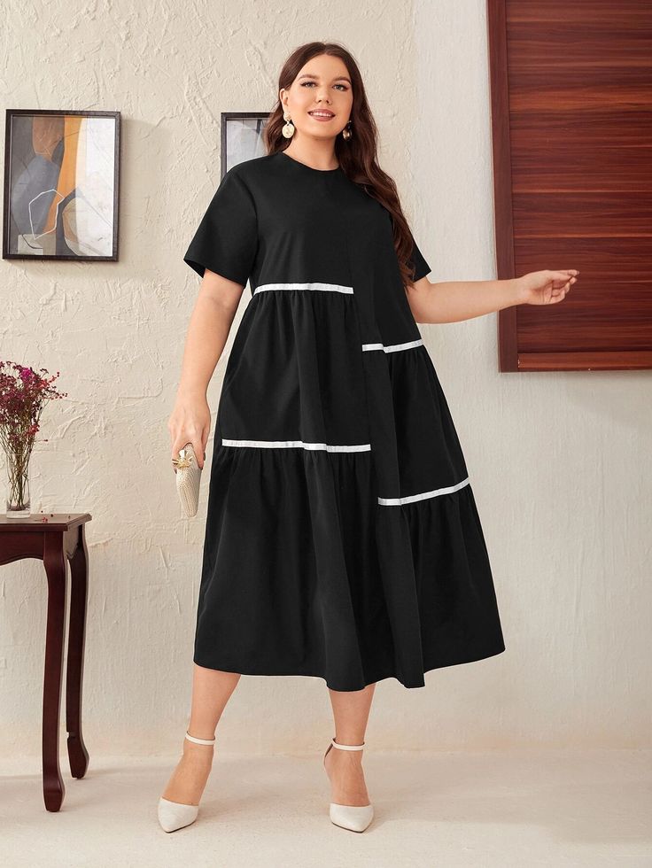a plussize model poses in a black midi dress with white trim. the dress is shortsleeved and tiered, with a casual and comfortable fit. the model is wearing white heels and holding a small clutch. the background is a white wall with a wooden floor.