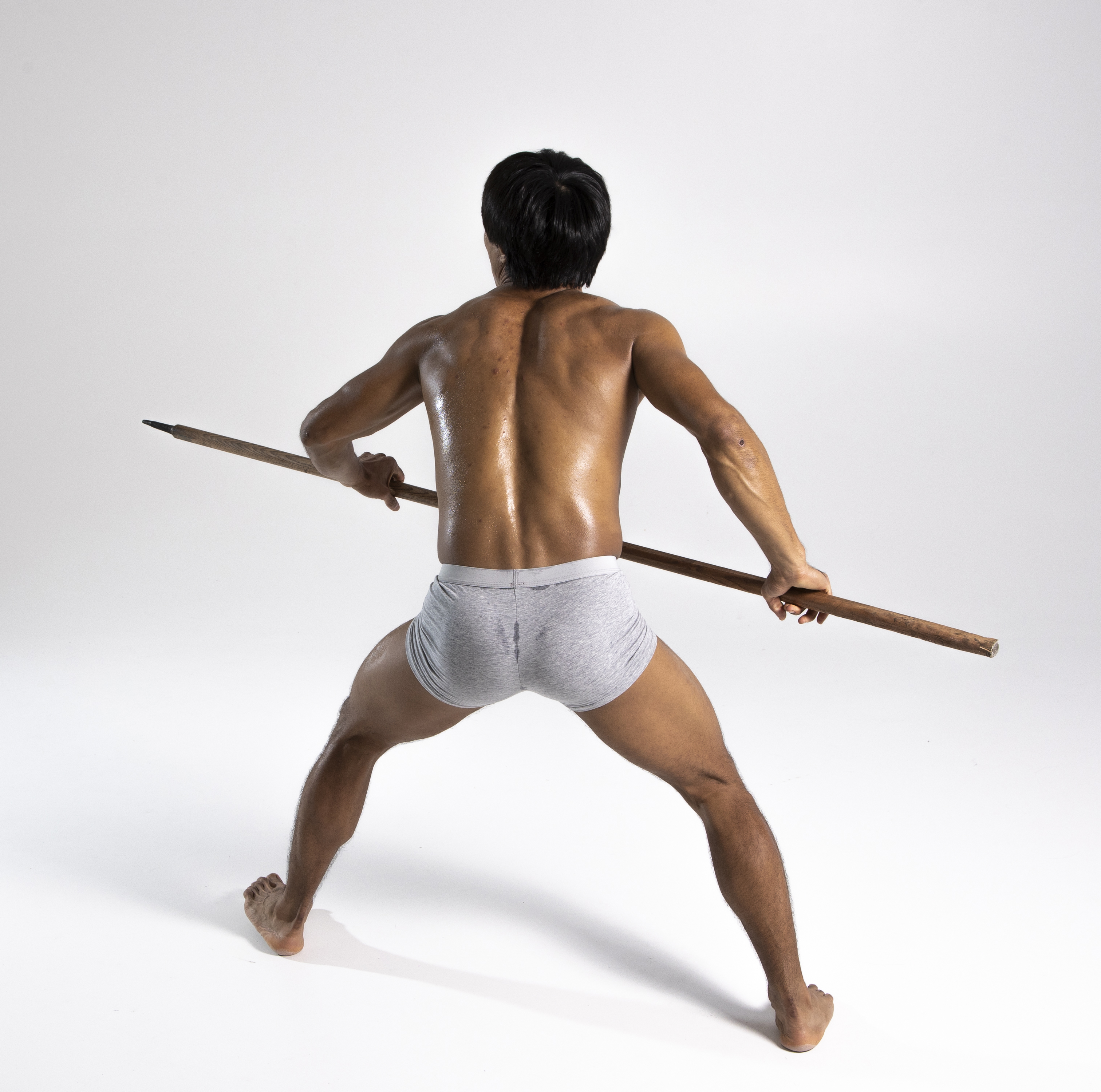 a man with a muscular build stands with his back to the viewer, holding a spear. he is shirtless, wearing only gray underwear. he appears to be in a defensive or aggressive stance, with his bare feet grounded. the background is a plain white studio setting.