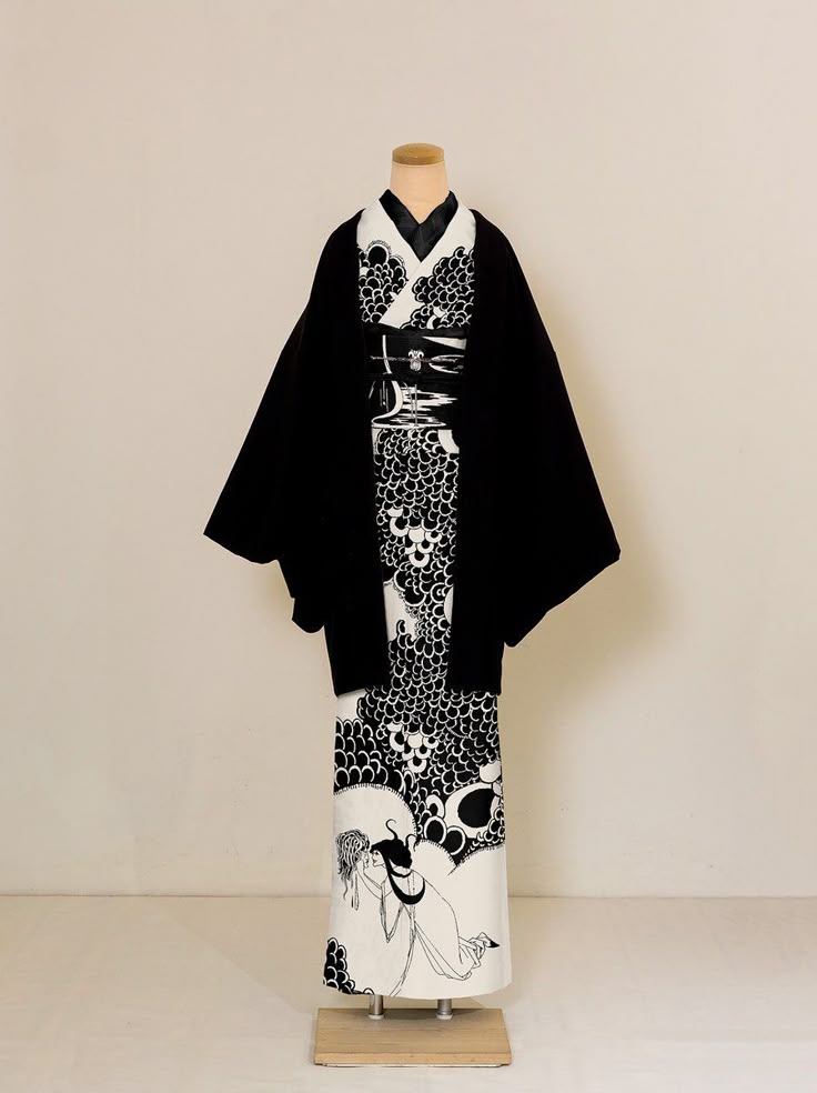 a black and white kimono with a black robe over it is displayed on a mannequin. the kimono has a intricate pattern and a black belt tied around the waist. the lower part of the kimono has an image of a person. the background is a plain, light colored wall.