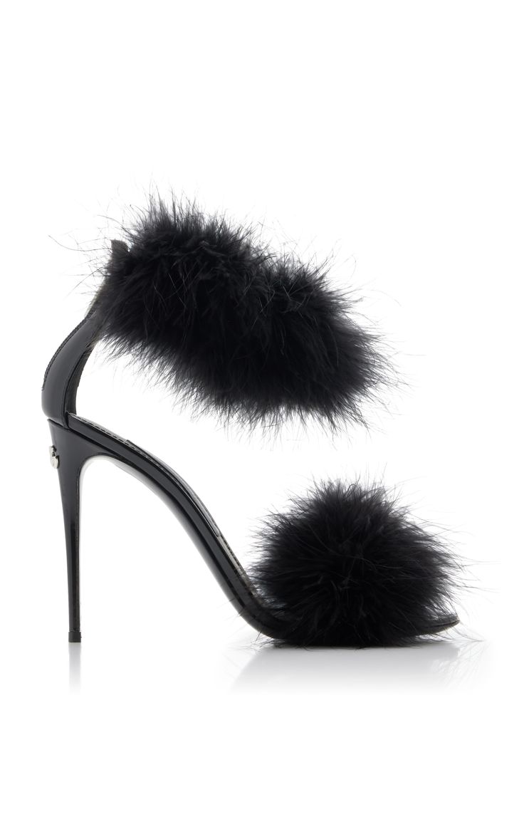 a pair of black highheeled sandals adorned with fluffy black feathers.  the feathers are arranged as two pompoms, one across the ankle strap and one at the toe. the shoes are made of patent leather with a stiletto heel.  the image shows the shoes from a side profile on a white background.