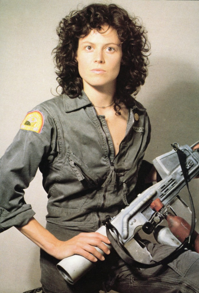a portrait of a woman with curly brunette hair, wearing a grey jumpsuit and holding a futuristic gun. she is looking at the camera with a serious expression on her face. this appears to be a still from a science fiction movie, possibly alien.