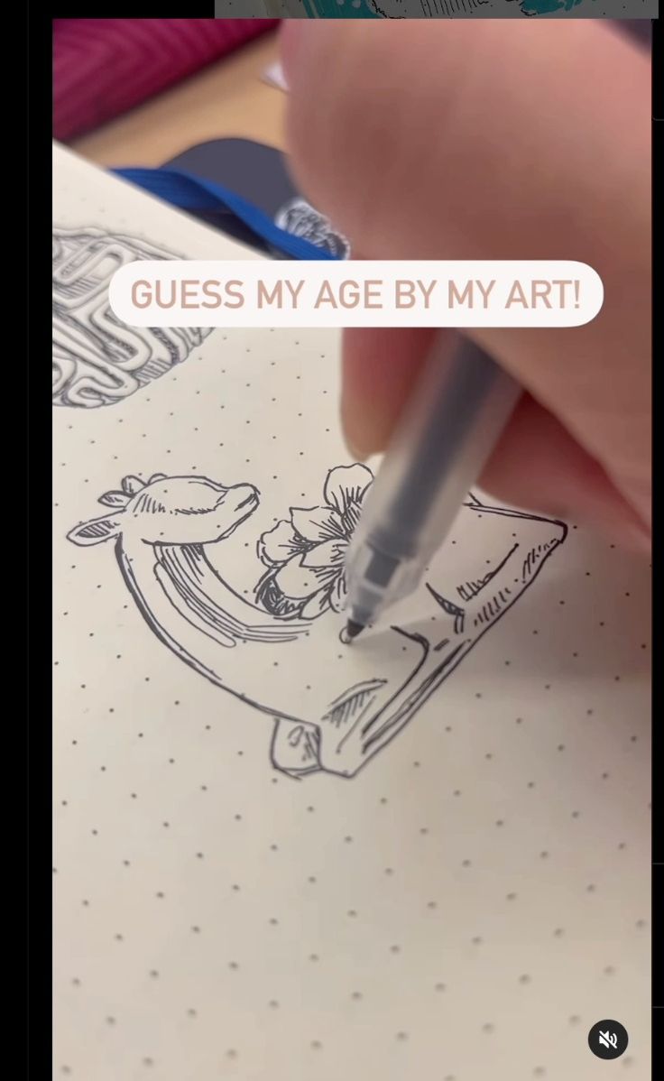 the image shows a closeup view of a person's hand drawing a sketch in a notebook. the drawing depicts a giraffe's head and neck entwined with a flower, possibly a whale's tail in the background. the notebook has dotted paper. the person's hand is holding a pen and is in the process of creating the artwork. a text overlay on the image asks viewers to "guess my age by my art!" the overall style of the drawing is simplistic and slightly whimsical. the image focuses on the drawing process and invites interaction from the viewer.